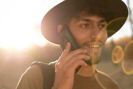Autumn evening. Sunset. Muslim young man standing in a park and talking on a cell phone, backlight. Handsome man uses a digital gadget.の写真素材