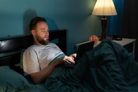 Handsome Caucasian man with laptop computer lying in bed at home at night. Technology, internet, communication and peopleの写真素材