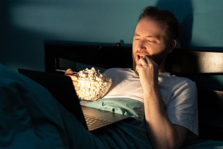 Handsome bearded man eating popcorn and watching film on laptop while resting in bed in bedroom at nightの写真素材