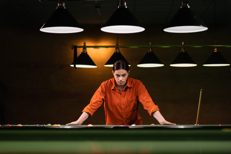 Pensive young woman at the billiard table holds a cue and thinks about the game