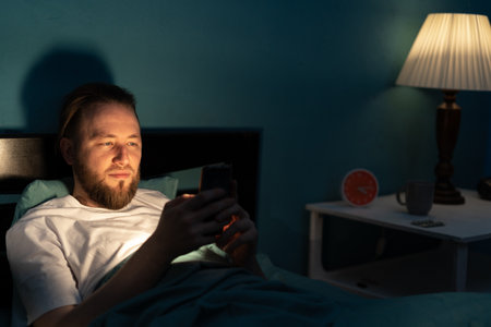 Millennial men uses smartphone in bed at night, suffering from insomnia and addiction. Alone man browsing social media, reading news and chatting at night.の写真素材