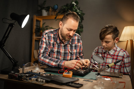 Young man and cute boy sitting together at home and testing motherboard of laptop. Father teaching son about repairing computer detailsの写真素材