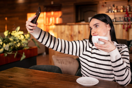 Gorgeous smiling brunette woman making a selfie while having a cup of coffee sitting in cafeの写真素材