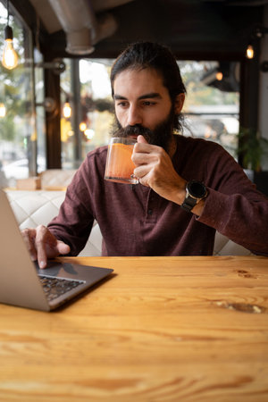 Portrait of bearded man working remotely on laptop computer in cafe interior, hipster guy drinking...