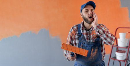 Painter man holding a paint roller standing in room shouting with mouth wide open to the sideの写真素材