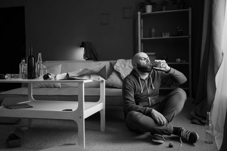 Male alcoholic with bottle of whiskey sitting on floor, black and white photo. Alcoholism, alcohol...
