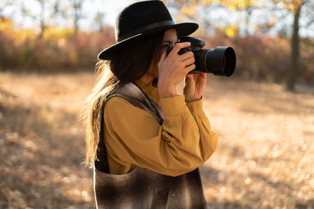 Cute woman in black hat with camera enjoys sunny weather in autumn park, photographer in yellow...
