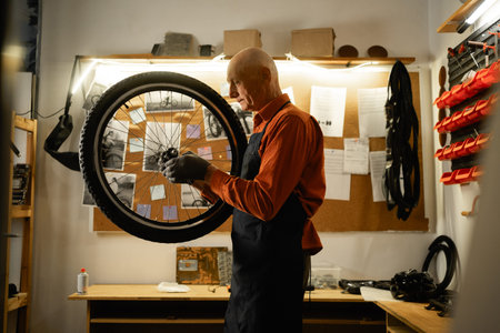Elderly male worker holding and repairing bicycle wheel while standing in authentic bicycle workshop.の写真素材