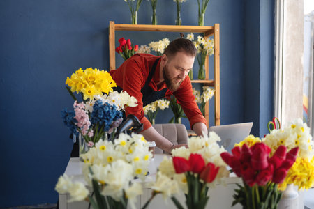 Male florist flower store owner working and doing inventory. Man typing on laptop at desk standing in floral shop. Business conceptの写真素材
