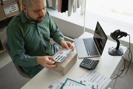 tired man sticks a barcode on parcel. Checks online with laptop. Small business.の写真素材