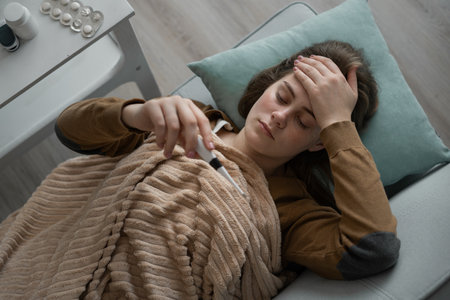 Sick young Caucasian woman with severe headache and fever measures temperature with thermometer, feels unwell, lying on sofa at home. Taking care of your health during the virus periodの写真素材