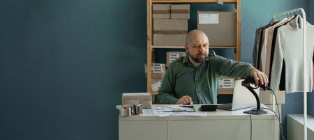 Tired man sitting, uses computer and barcode scanner to record customers online data. Startup...