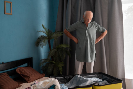 old man traveler packing suitcase with clothes stands thoughtfully near suitcase and luggage at home.の写真素材