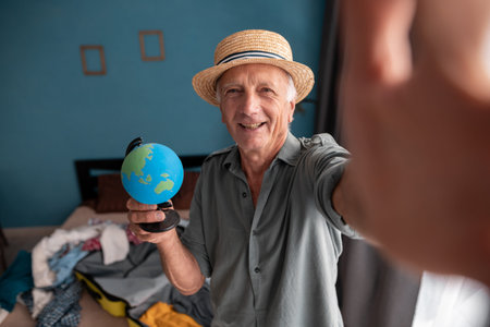 Happy elderly traveler man taking selfie portrait while preparing travel suitcase on summer vacation...