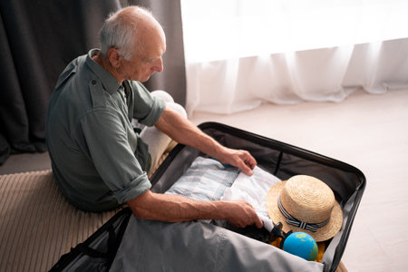 Travel concept. Retirement man preparing packing luggage bag for summer vacation trip sitting on bed at home, pack clothes into suitcase for a new journey.の写真素材