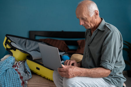 Travel, summer and holiday concept. Senior man prepare things into luggage for trip at home. Grandfather using computer to planning travel trip and booking tickets online.の写真素材