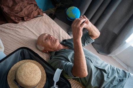 Senior old active man travelling, using globe lying on the bed check the route, going on a holiday, voyage with luggageの写真素材