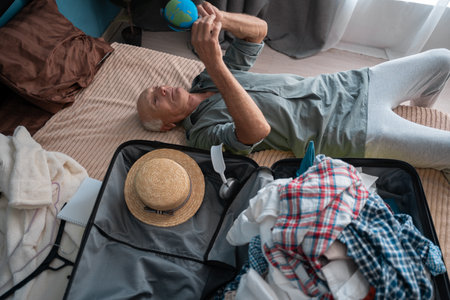 Elderly man packing his travel essentials on suitcase, getting ready to start vacation. Top view Retired traveler lying on the bed dreaming adventure in a tourist destinationの写真素材