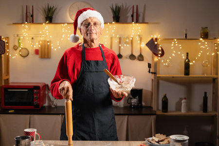 Senior man prepares gingerbread cookies in kitchen on Christmas Eve holding rolling pin and dough bowl standing against background of lights creating a cozy festive holiday atmosphere.の写真素材