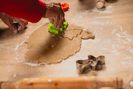 Close-up of senior hands using green cutter to create Christmas tree shapes from dough. Festive holiday cookie preparation in a cozy kitchen with flour and rolling pin.の写真素材