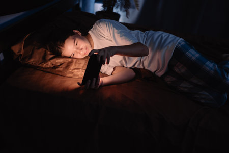 Teenage girl playing online game using mobile phone while lying in bed at night. Concept of insomnia and addictionの写真素材