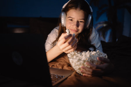 Teen girl wear headphones with long dark hair lying on bed watching interesting movie and eating popcorn on laptop at nightの写真素材