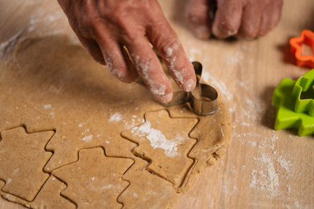 Person cutting gingerbread tree cookie from dough, Christmas holiday advent. Cooking process gingerbreads and cookies with plastic cutter on background of festive lights and rustic tableの写真素材