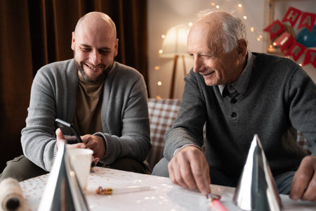 Happy people and family concept. Old senior man with his son celebrating birthday looking in smartphone happily. Young man near his elderly father on the sofa.の写真素材