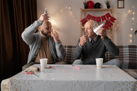 Old dad and his middle-aged son puts party hat in home for birthday celebration. People, family and bonding in special eventの写真素材
