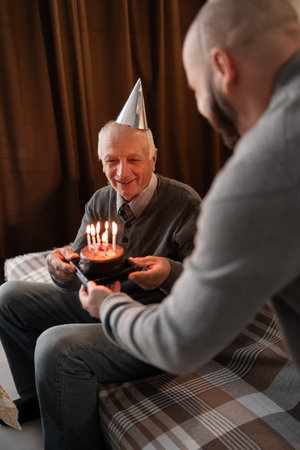 Elderly father with adult son celebrates birthday at home. Man present him with a cake, creating a...