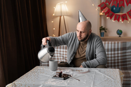 Sad depressed bearded man in party hat sitting alone on sofa celebrating his birthday and pouring tea in cup birthdayの写真素材