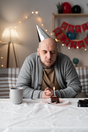 Man celebrating birthday alone sitting on sofa making wish and blowing out candle on piece of cakeの写真素材