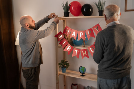 An adult man and his elderly father decorate a room with flags with an inscription standing near a bookshelf in the living room of a house.の写真素材