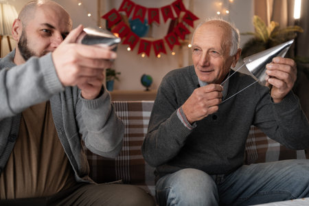 Senior father and adult son preparing to celebrate birthday or anniversary putting party hats on heads sitting on the sofa in living roomの写真素材