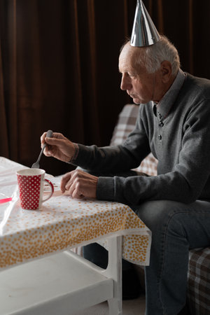 Lonely elderly man celebrating birthday alone eating cake and drinking tea at home on sofaの写真素材