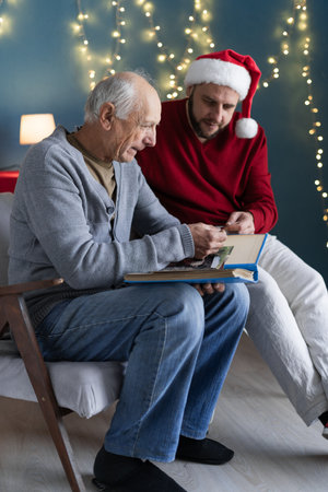 Two people senior man and his son looking at a family photo smile sitting at home on armchair in Christmas. memories and nostalgia with their familyの写真素材