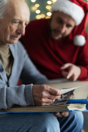 family with senior father and adult son enjoying free time at home on Christmas. Parent and child sitting on comfy armchair, looking through holiday photo album togetherの写真素材