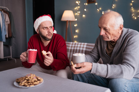 Father and son share holiday moment drinking hot cocoa and eating cookies, surrounded by festive lights.の写真素材