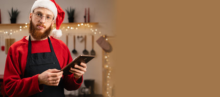 Bearded Man in Santa hat making dough while watching online cooking course via tablet in decorated kitchenの写真素材