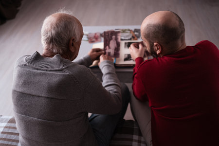Elderly man looking at old photo album with his son or friend sitting on sofa at homeの写真素材