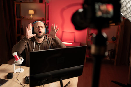 A professional gamer-blogger man shoots a video blog while playing a video game at night. the cyber athlete vlogs, folds his hands near his mouth and shouts something into the cameraの写真素材