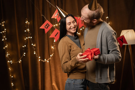 Young man in party hat hugs and kisses his beloved wife giving gift for her. Concept of gifts for birthdayの写真素材