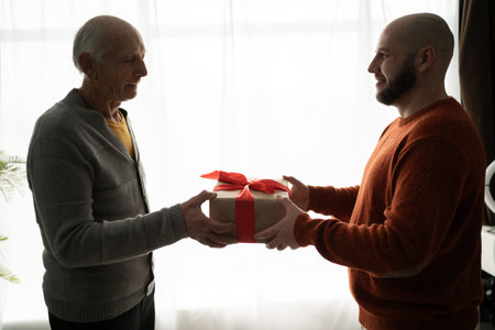 Son giving gift decorated with red bow to retired father standing near window in living room, side...