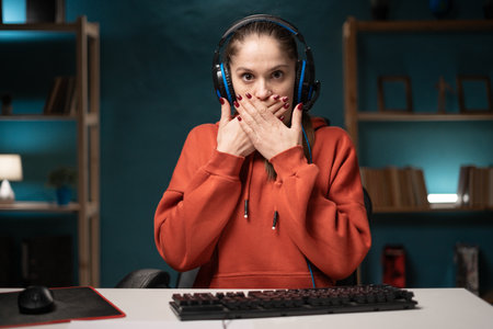 Shocked woman gamer play game. Female gaming player wearing headset with amazed face expression covering her mouth while broadcast live streaming online game at home. POV. Webcam view.の写真素材
