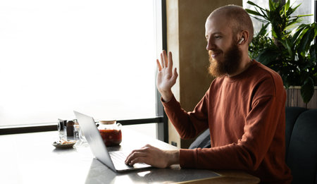 cheerful redhead man millennial with beard talking online on laptop in cafe, having remote...