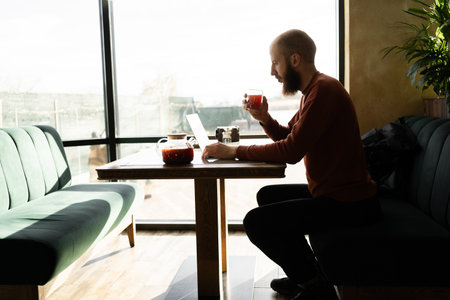 Bearded hipster man work or study on laptop computer drink tea sitting in cafe indoors. Freelance businessの写真素材