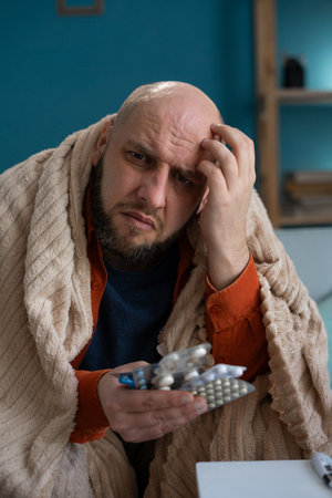 Sick man wrapped in blanket holding pillsの写真素材