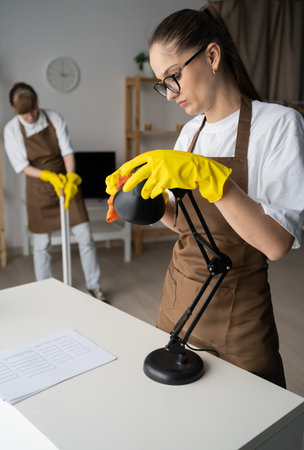 Female professional cleaners wiping desk lamp in living room. team of janitors from cleaning service workingの写真素材