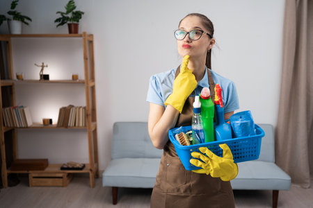 portrait of woman with cleaning basket for home service. Female worker holding product for housekeeping in cleaner uniform with dreamy look, thinkingの写真素材