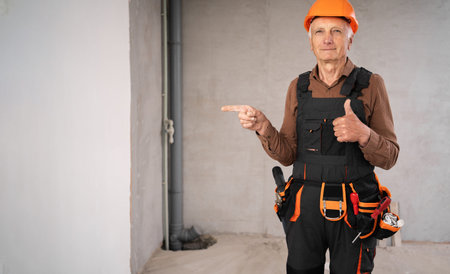 Senior builder on construction site standing wearing safety helmet pointing thumb-up and finger on...